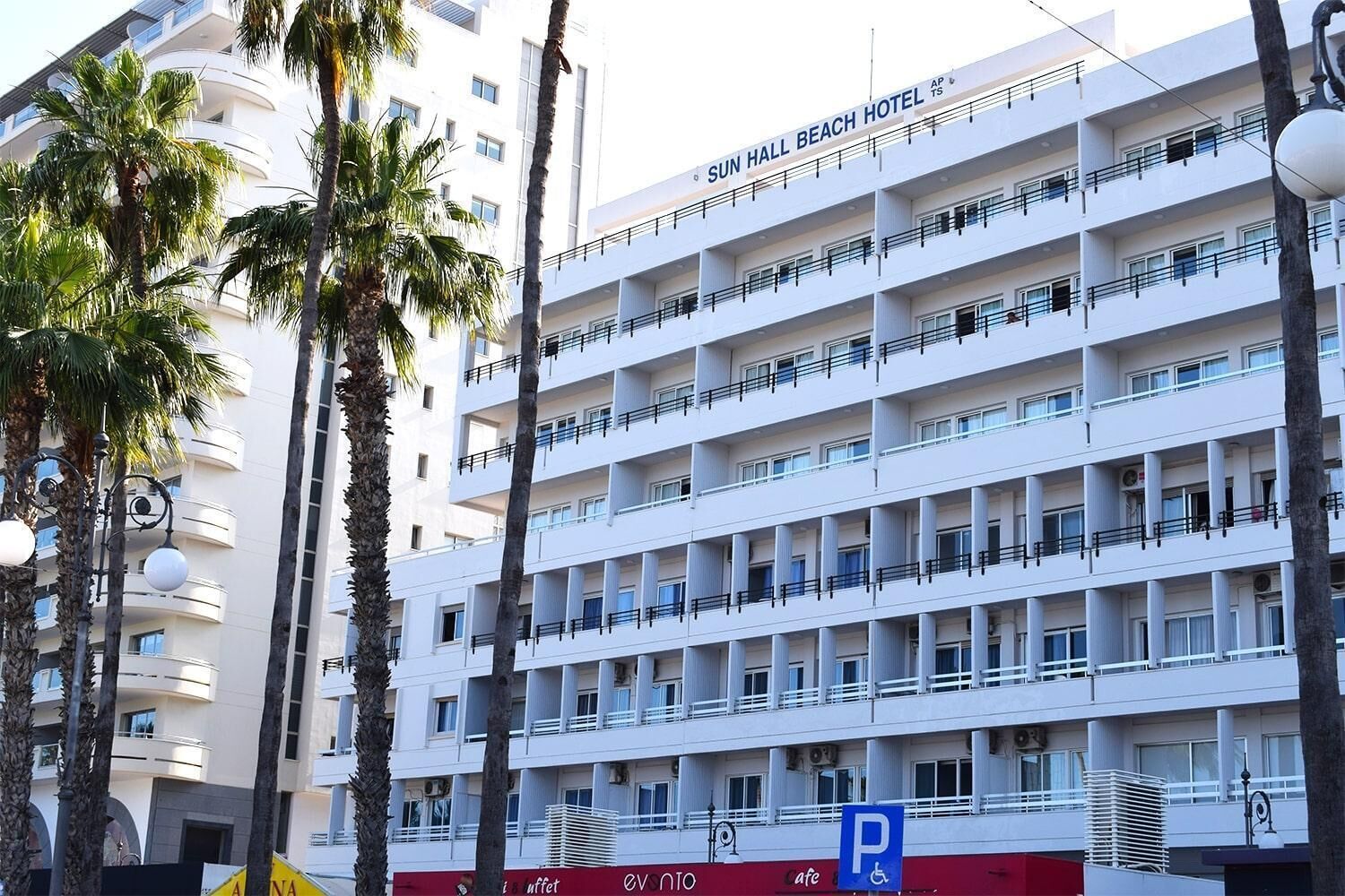Un hotel de varios pisos con balcones, palmeras y un cartel de estacionamiento.