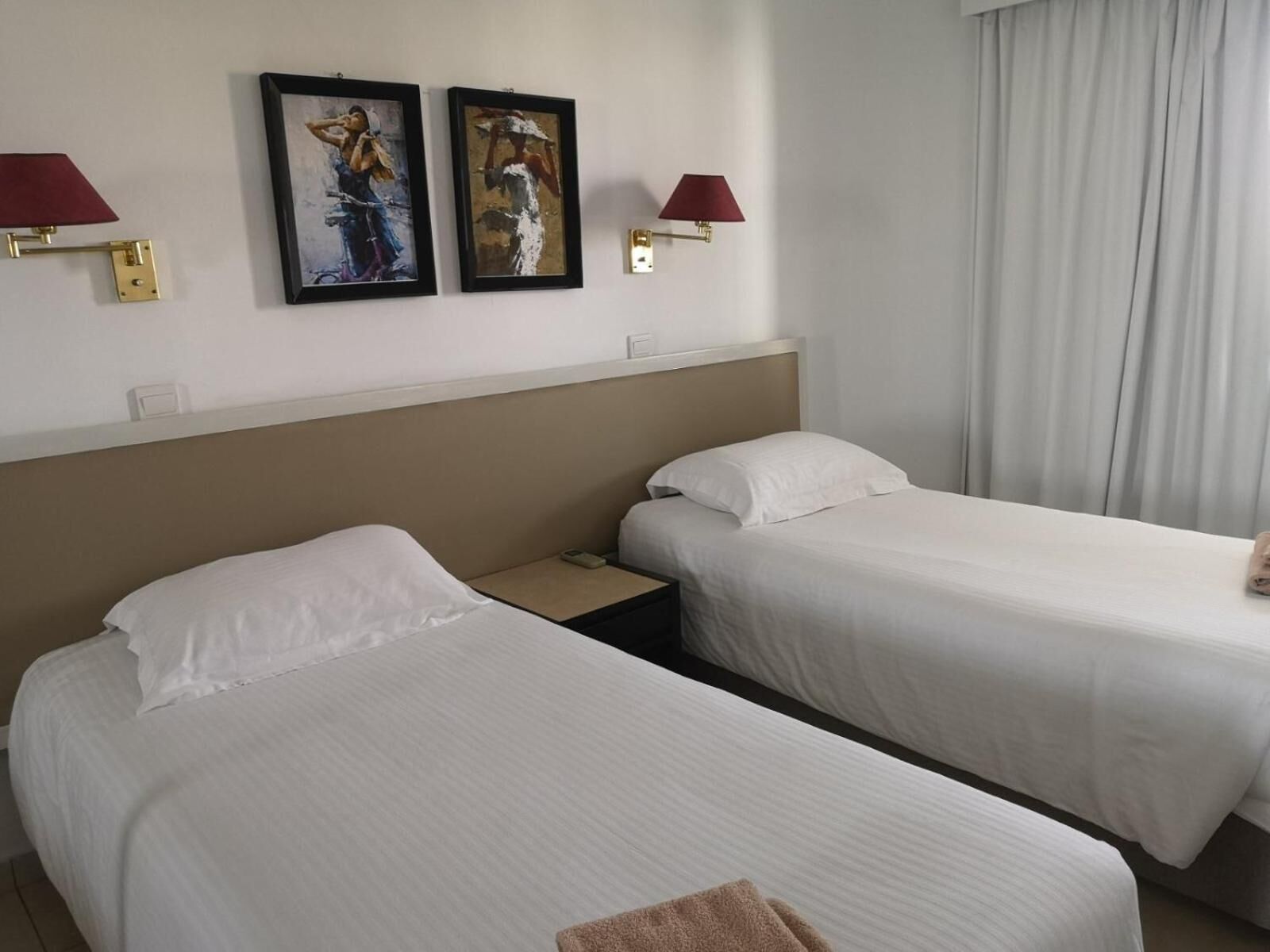 A hotel room with two single beds, a nightstand, a lamp, and two framed artworks on the wall.