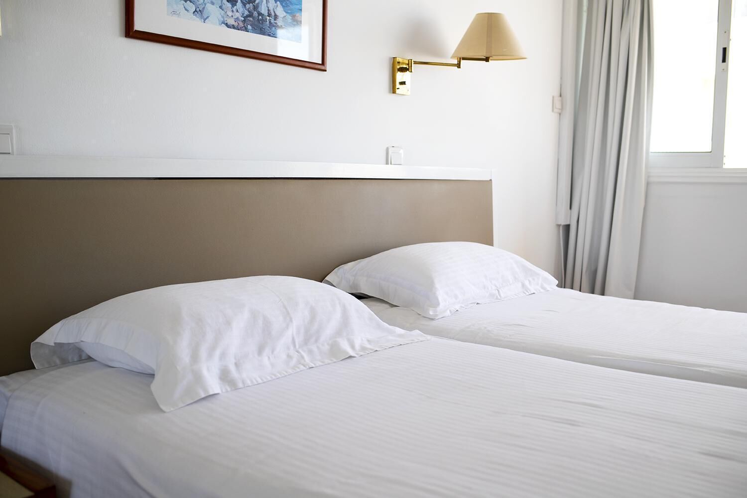 A hotel room with two single beds, a headboard, a wall-mounted lamp, and a window with curtains.