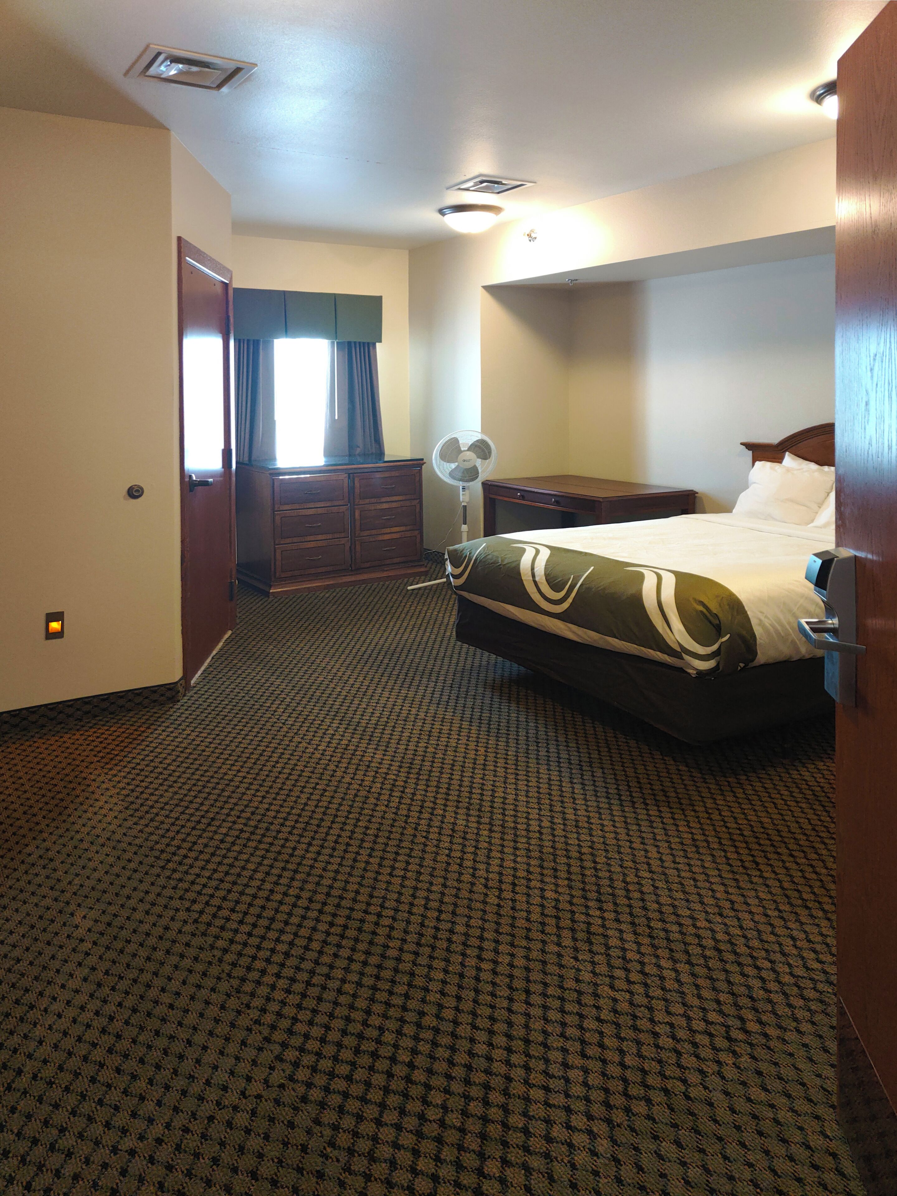 Una camera d'albergo con un letto, una cassettiera, una scrivania, una ventola e una finestra con tende.