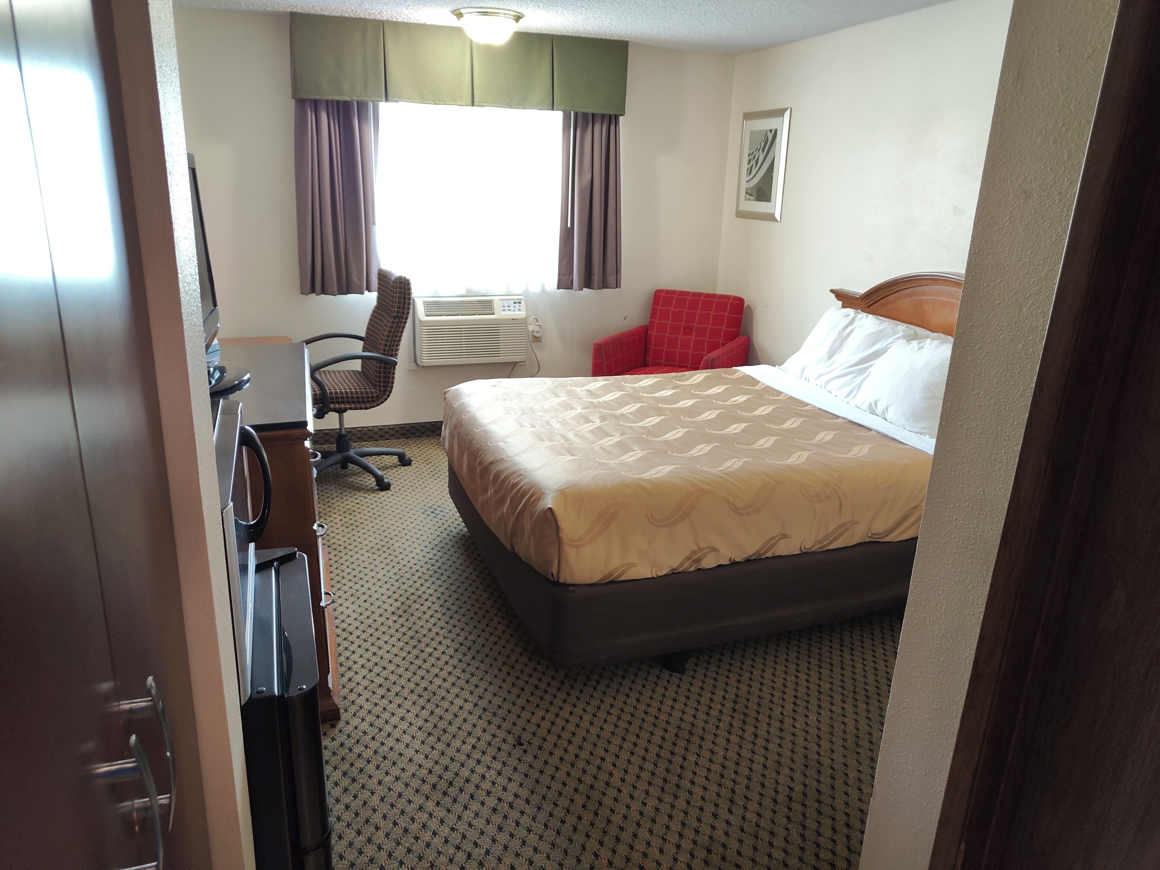 A hotel room with a bed, a desk, a chair, an air conditioning unit, and a window with curtains.