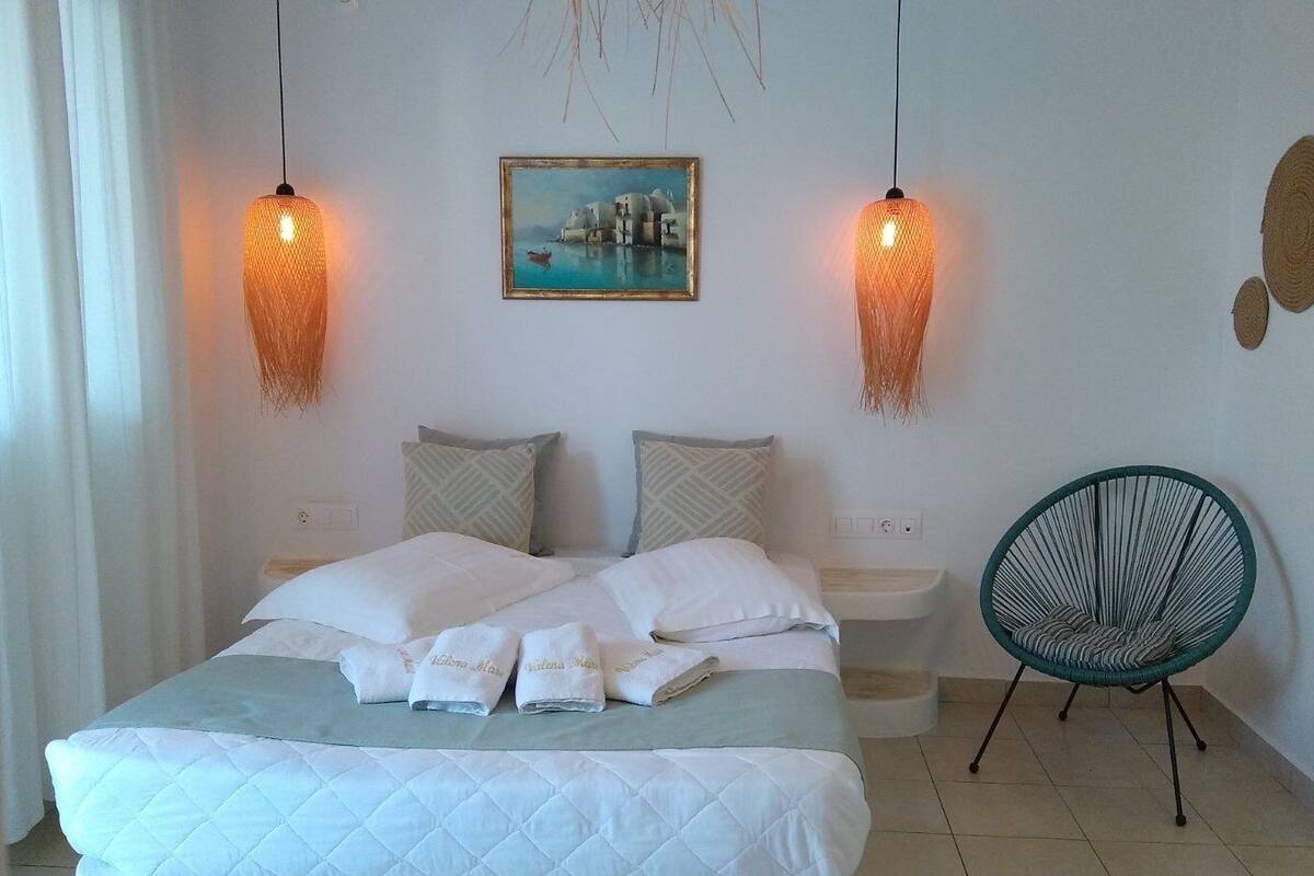 Uma cama bem arrumada com lençóis e toalhas brancas, duas mesas de cabeceira, uma cadeira e um quadro com uma paisagem costeira na parede.