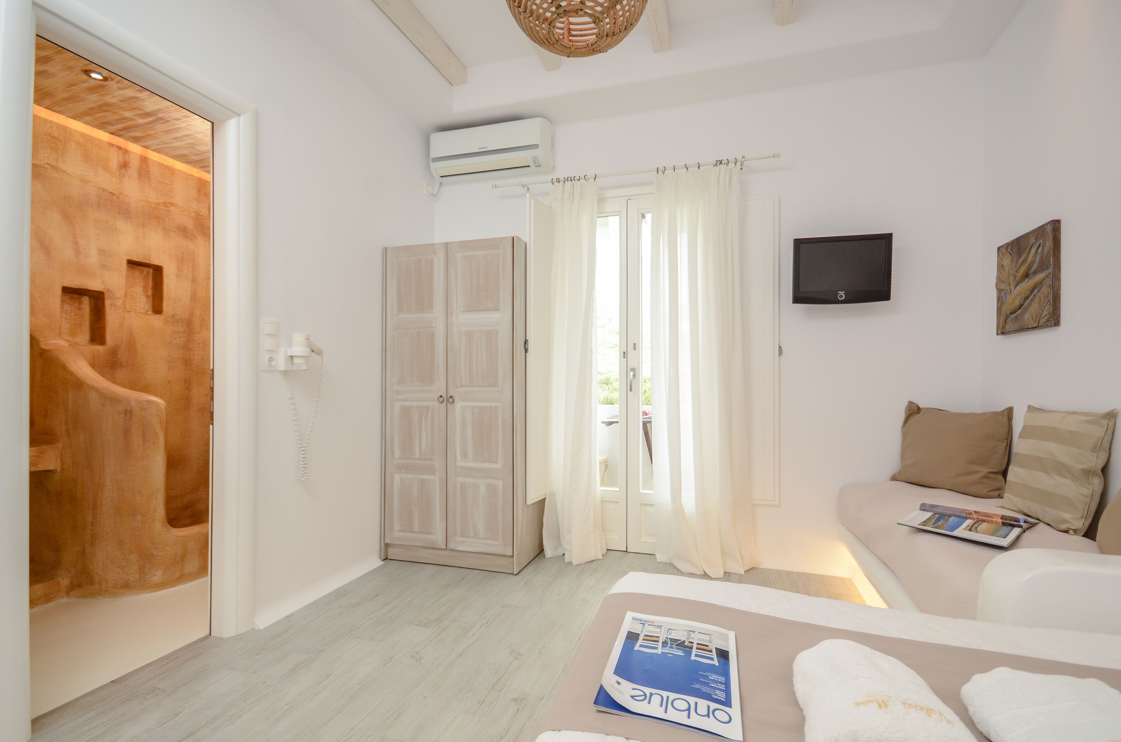 A room with a wooden door, a white sofa with pillows, a flat-screen TV, and a magazine on the table.