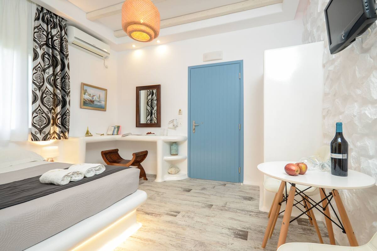 Quarto de hotel moderno com cama, escrivaninha, cadeira e uma pequena mesa com uma garrafa e maçãs.