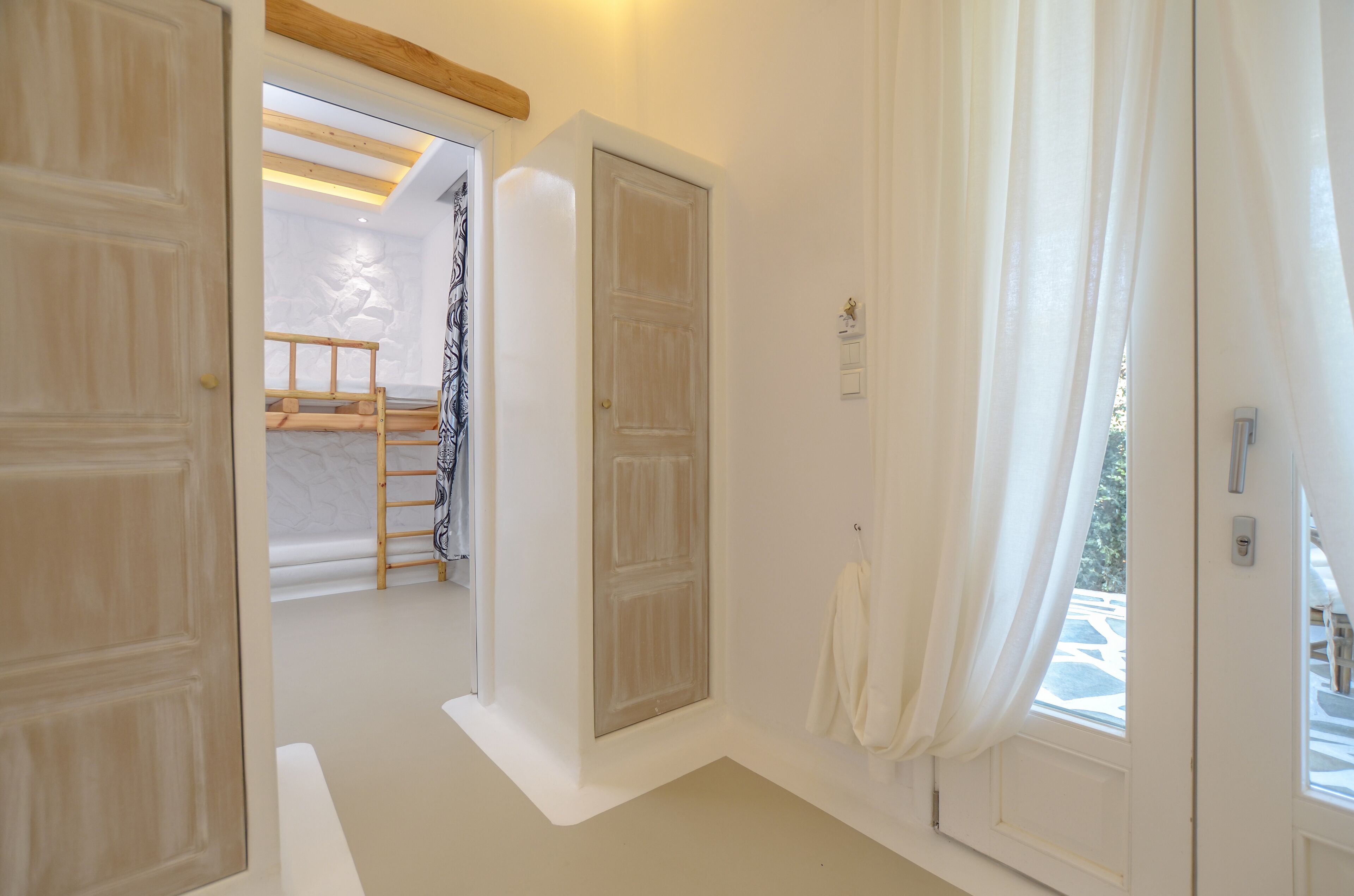A hallway with white walls, wooden doors, and a window with white curtains. A wooden ladder is visible in the background.