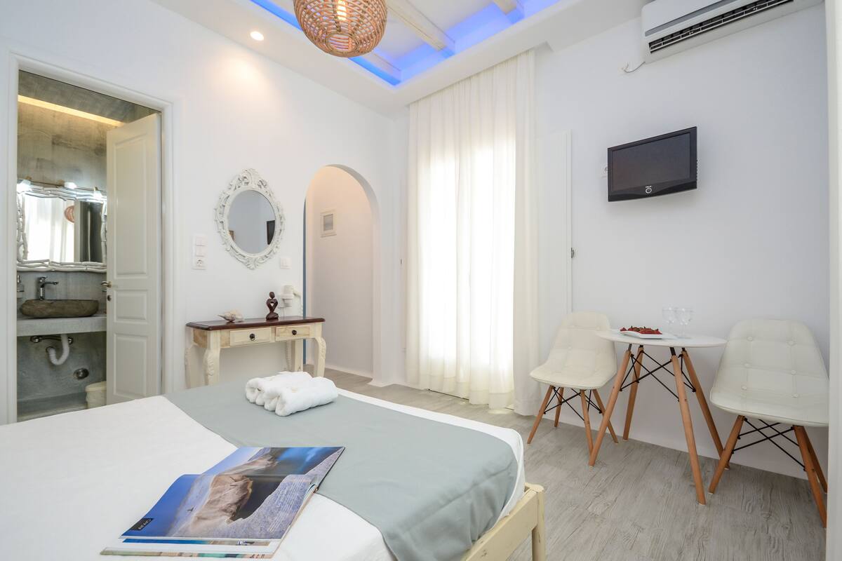 Quarto de hotel com cama, escrivaninha, duas cadeiras, televisão, espelho e banheiro com pia e chuveiro.