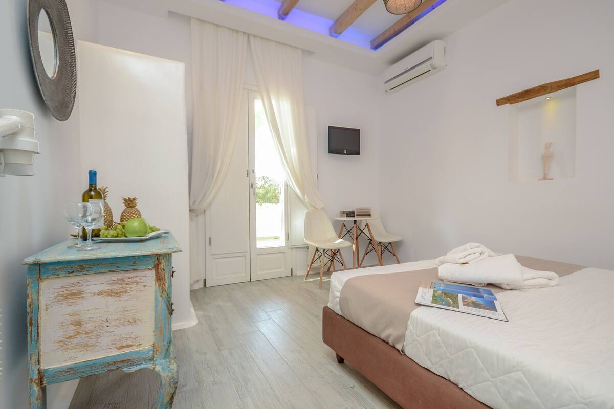 Um quarto com uma cama, uma mesinha com uma garrafa e uvas, uma cadeira, um espelho e uma televisão na parede.