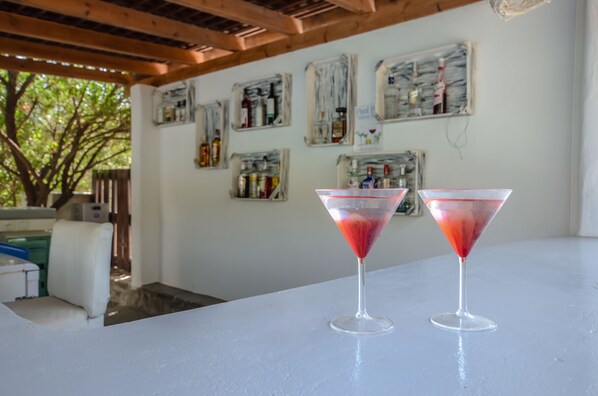 Deux verres à martini sur une table blanche, sous un plafond en bois et devant un mur orné de bouteilles d’alcool encadrées.