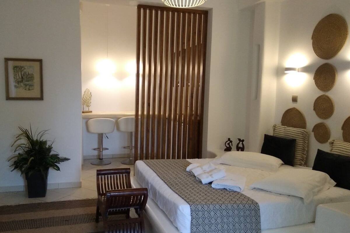 Quarto de hotel moderno com cama, escrivaninha, cadeira, planta em vaso e banheiro com espelho e porta-toalhas.
