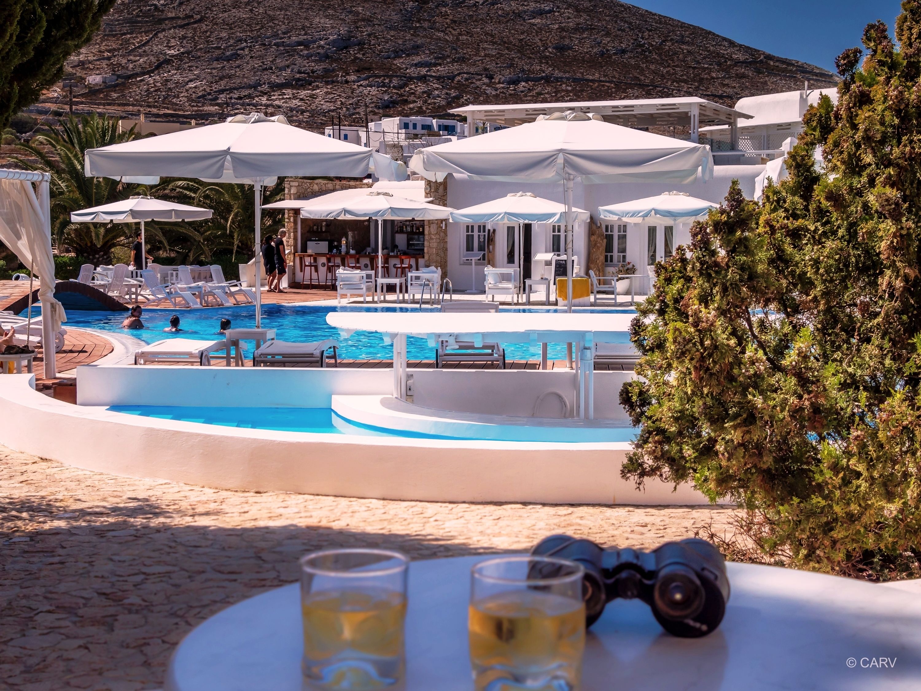 Zona de piscina con sillas de descanso y sombrillas, un bar y vistas a un paisaje montañoso.