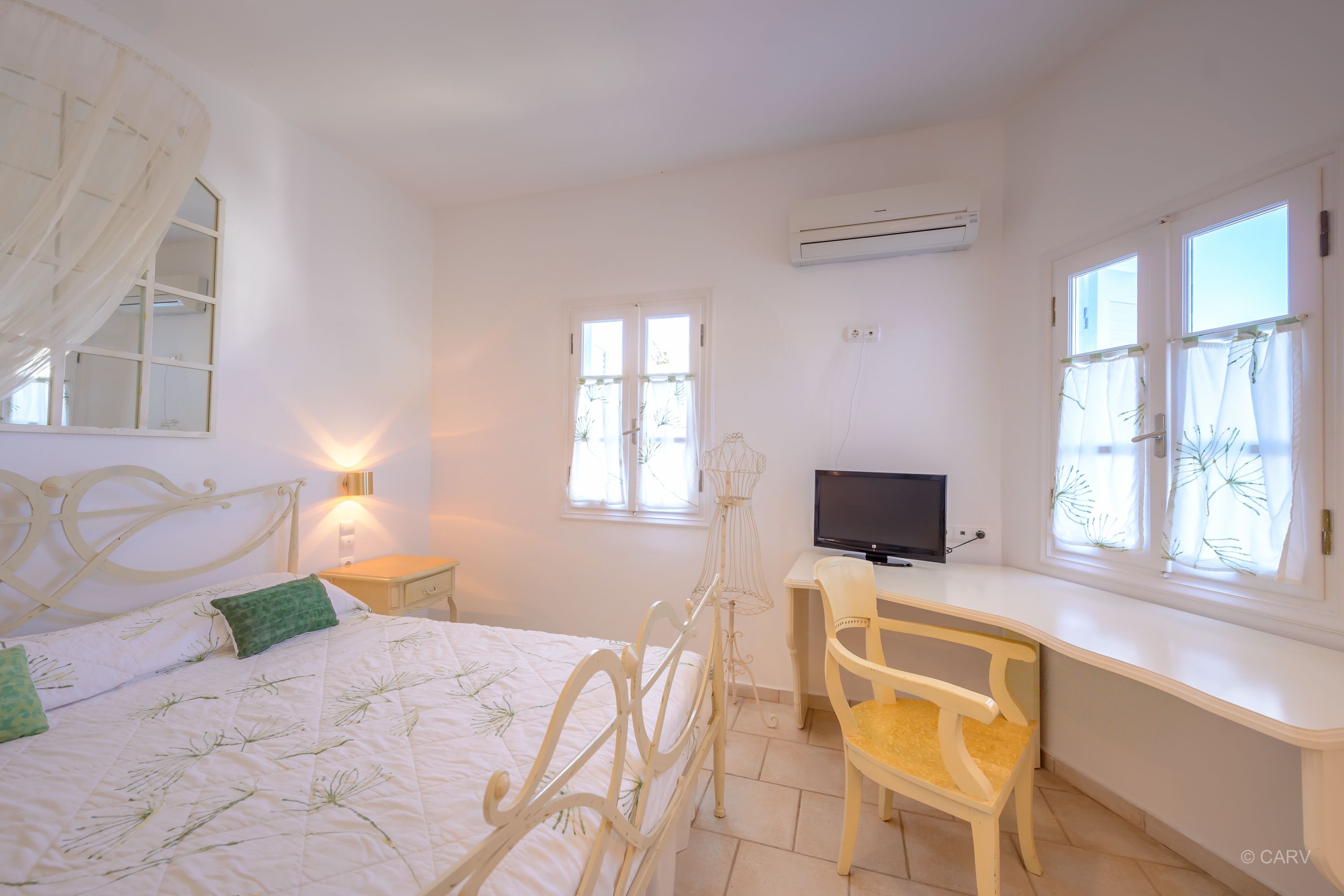 Un dormitorio con cama, mesita de noche, televisor, escritorio, silla y ventanas.