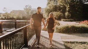 A couple walking hand in hand on a wooden boardwalk with a metal railing, surrounded by greenery and flowers. - Hôtel Château Joliette (Joliette)