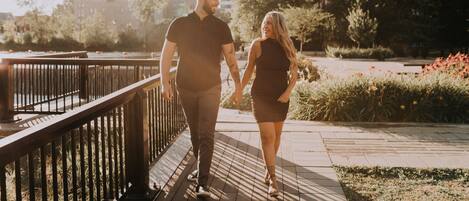 A couple walking hand in hand on a wooden boardwalk with a metal railing, surrounded by greenery and flowers.