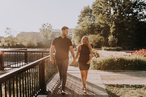 Una pareja caminando de la mano por una pasarela de madera con barandilla de metal, rodeada de follaje y flores.