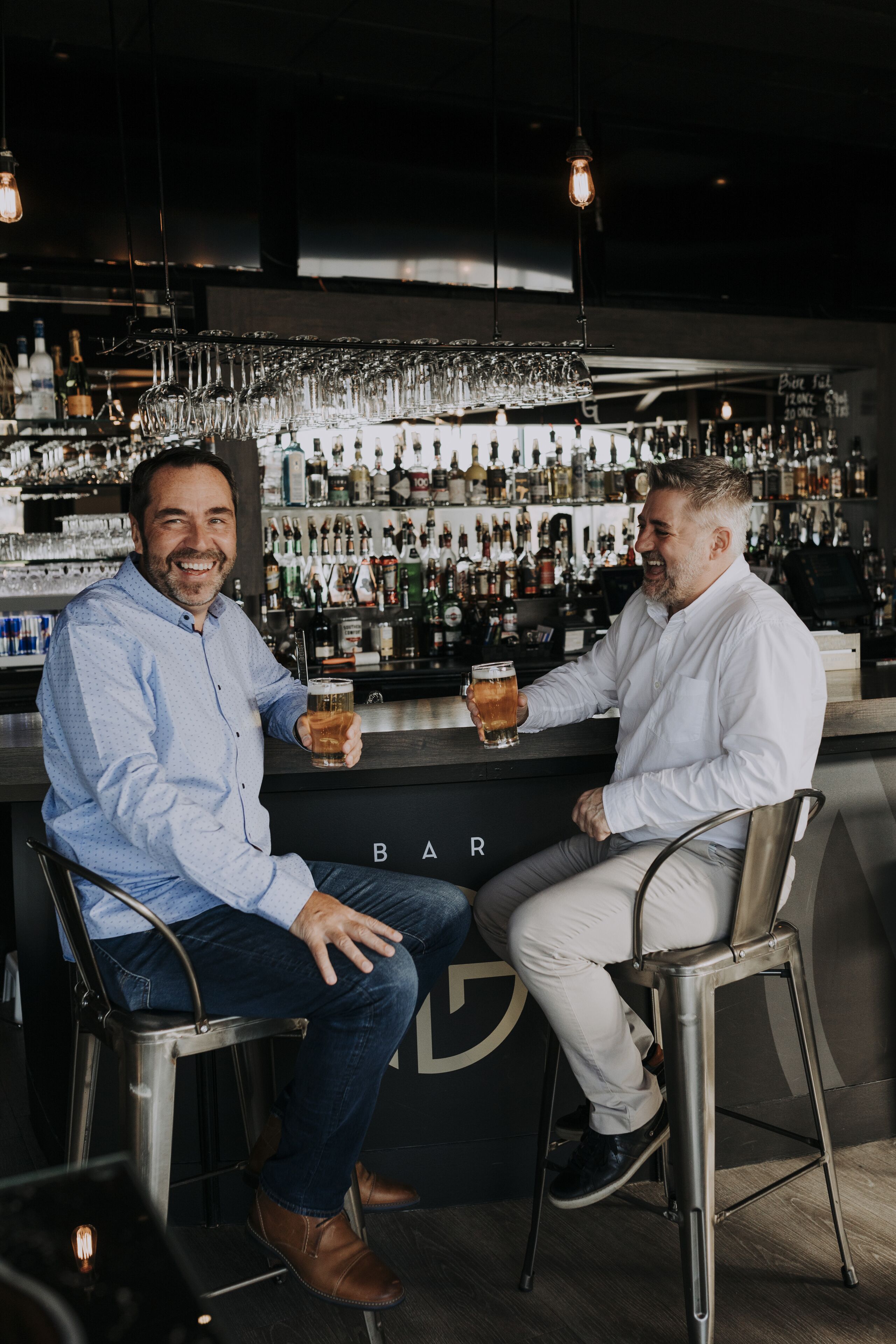 Two men seated at a bar with a variety of bottles and glasses in the background.