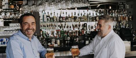 Two men seated at a bar with a variety of bottles and glasses in the background.