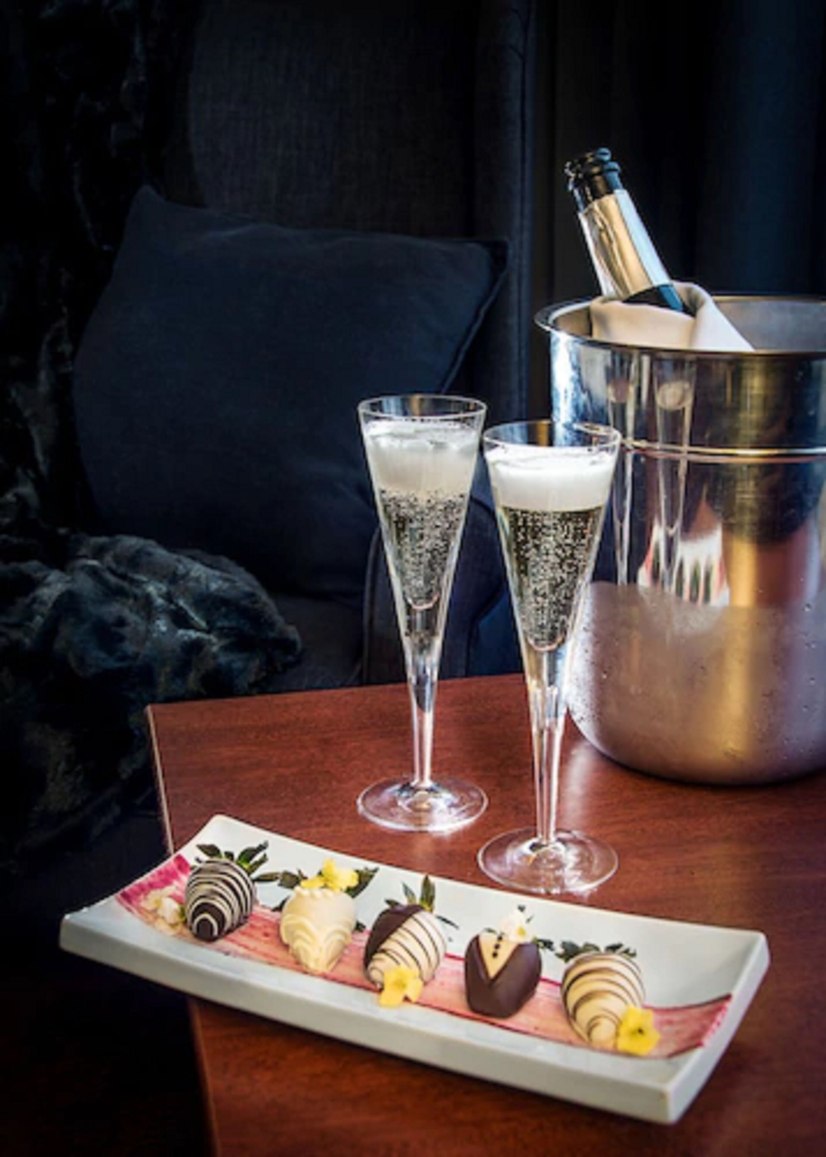 A table with two champagne flutes, a bucket of champagne, and a plate of chocolate-covered strawberries.