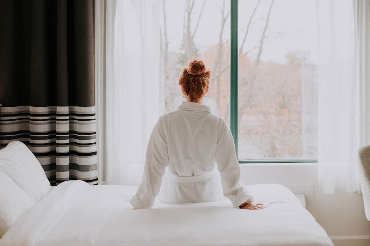 A person standing at the foot of a bed facing a window with sheer curtains.