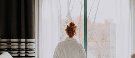 A person standing at the foot of a bed facing a window with sheer curtains.