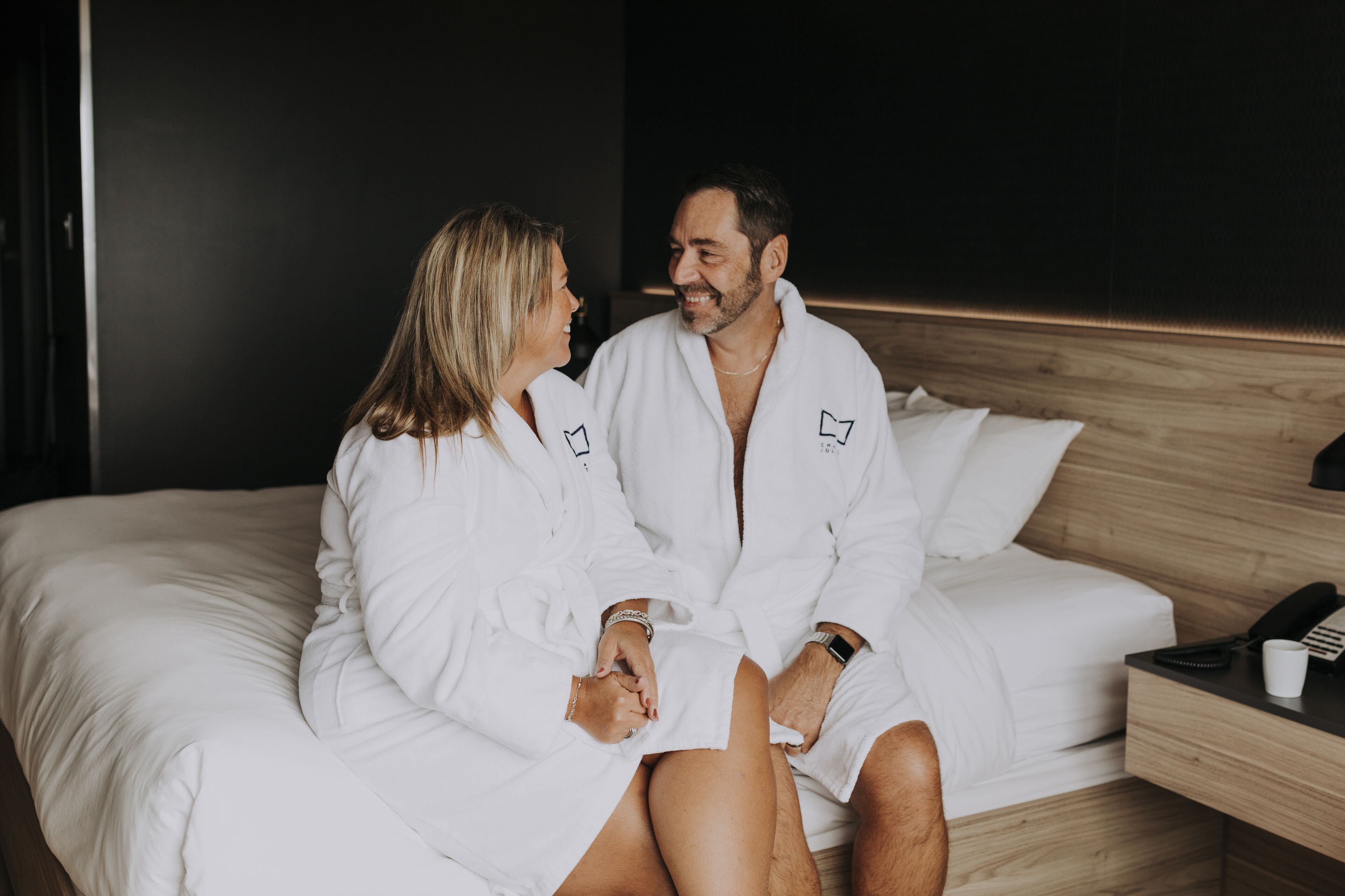 A couple in white robes sitting on a bed with a wooden headboard, a bedside table with a phone and a cup, and a dark wall in the background.