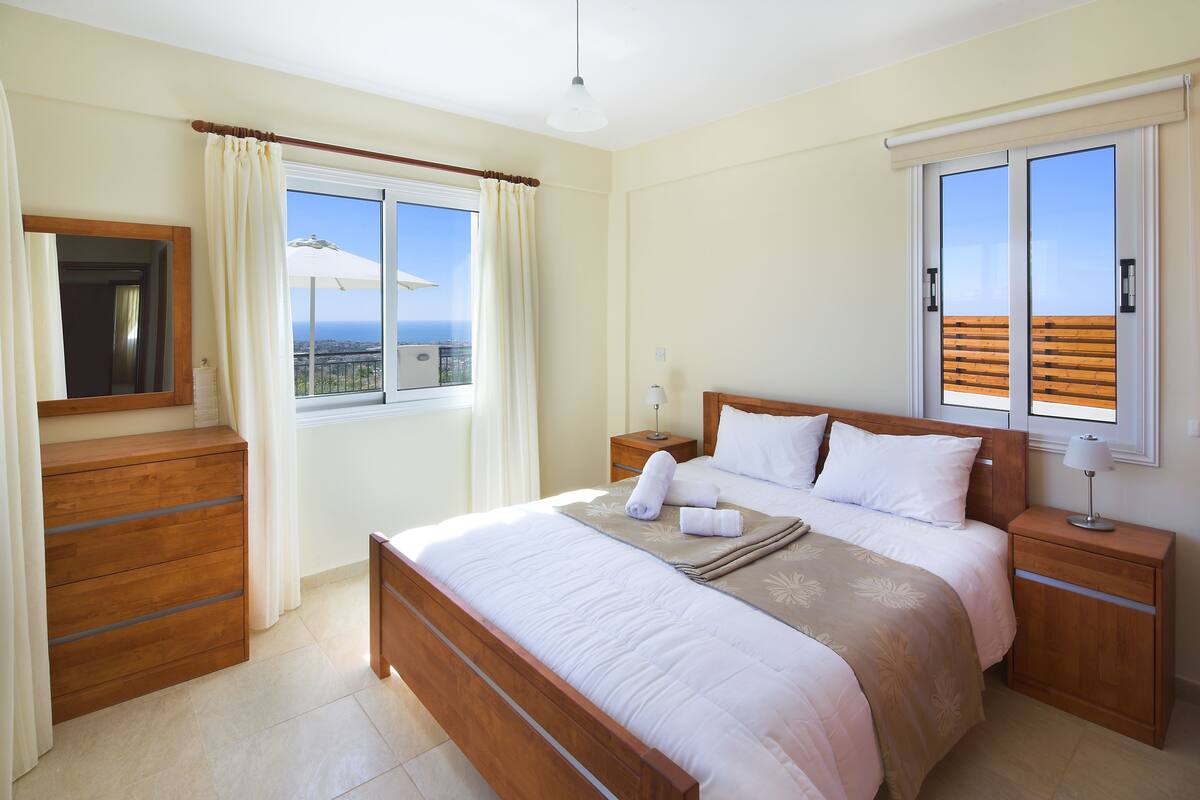 A bedroom with a bed, nightstands, a wooden dresser, a window with curtains, and a view of the ocean.