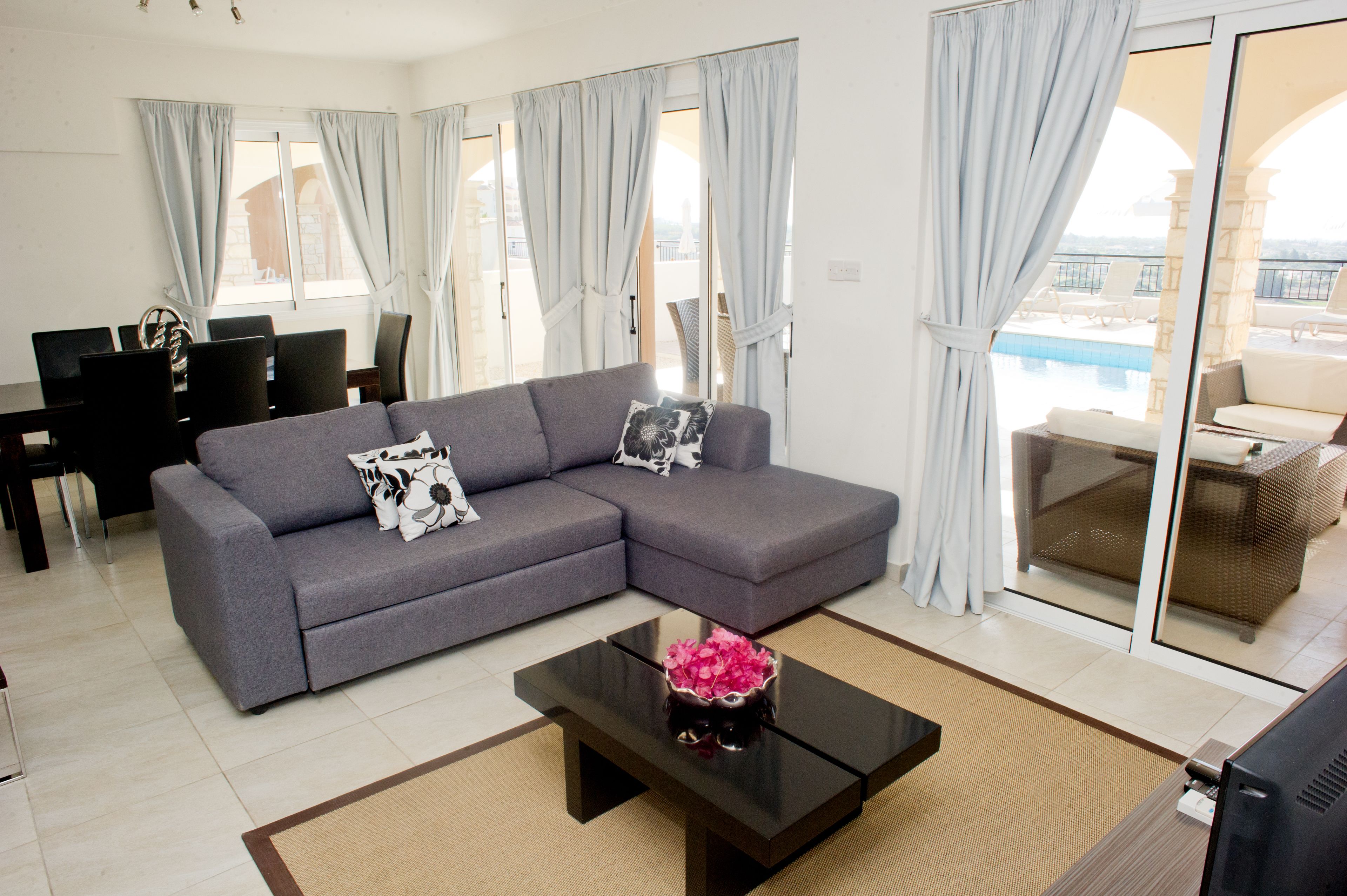 A modern living room with a grey sectional sofa, a black coffee table with a pink flower vase, and a dining area with a dark wooden table and chairs.