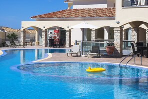 A swimming pool with lounge chairs and umbrellas, surrounded by stone pillars and a tiled roof.
