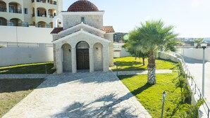 Església de pedra amb cúpula i entrada apuntada, envoltada per un jardí ben cuidat i una palmera.
