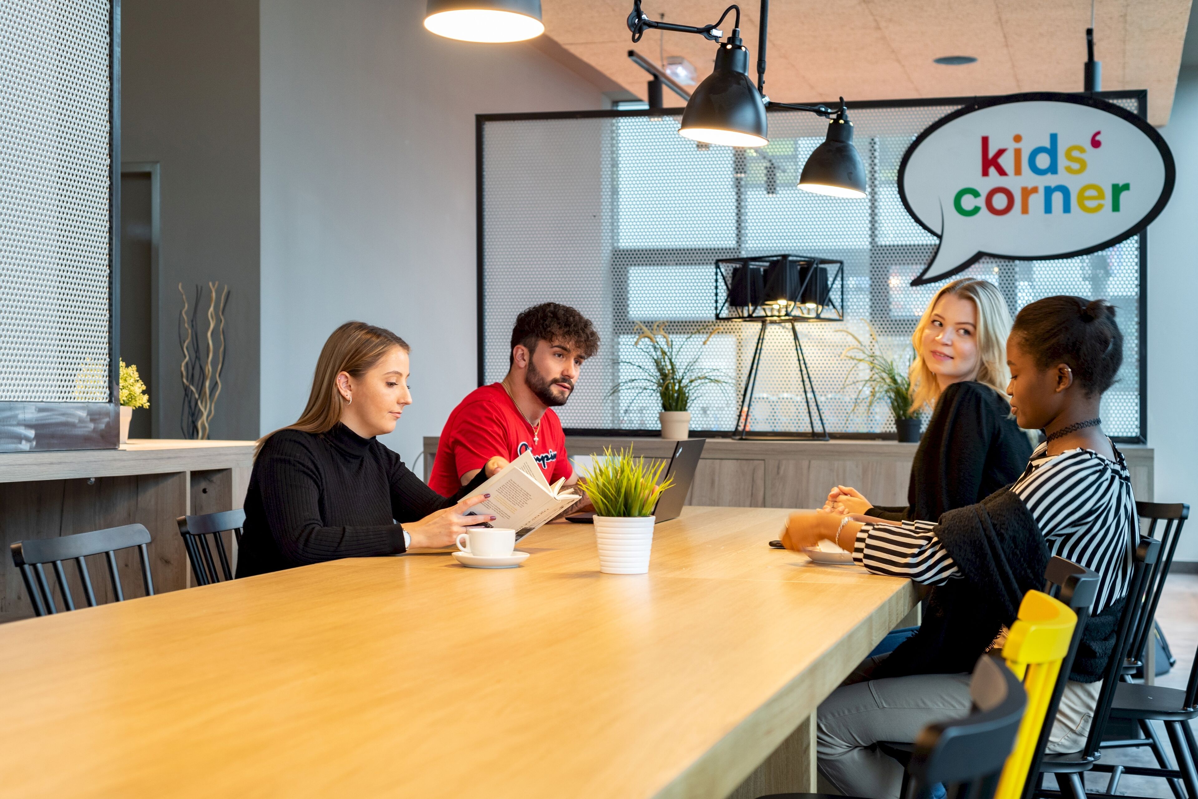 A group of people seated around a wooden table in a modern indoor setting with a sign reading "kids' corner".