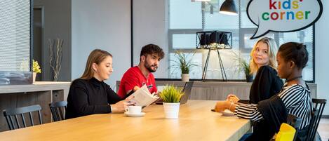 A group of people seated around a wooden table in a modern indoor setting with a sign reading "kids' corner".
