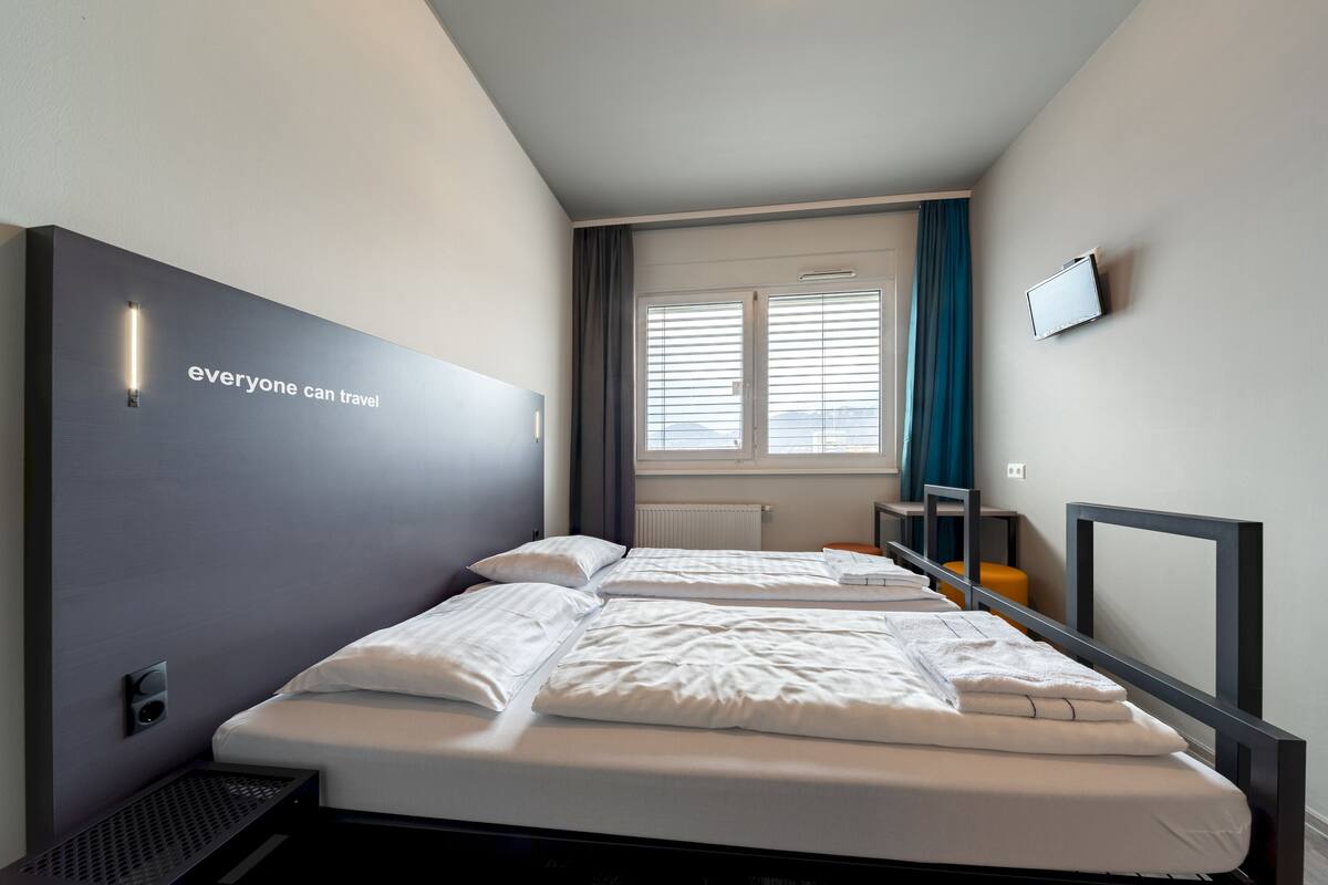 A modern hotel room with a large bed, a TV mounted on the wall, and a window with blinds.