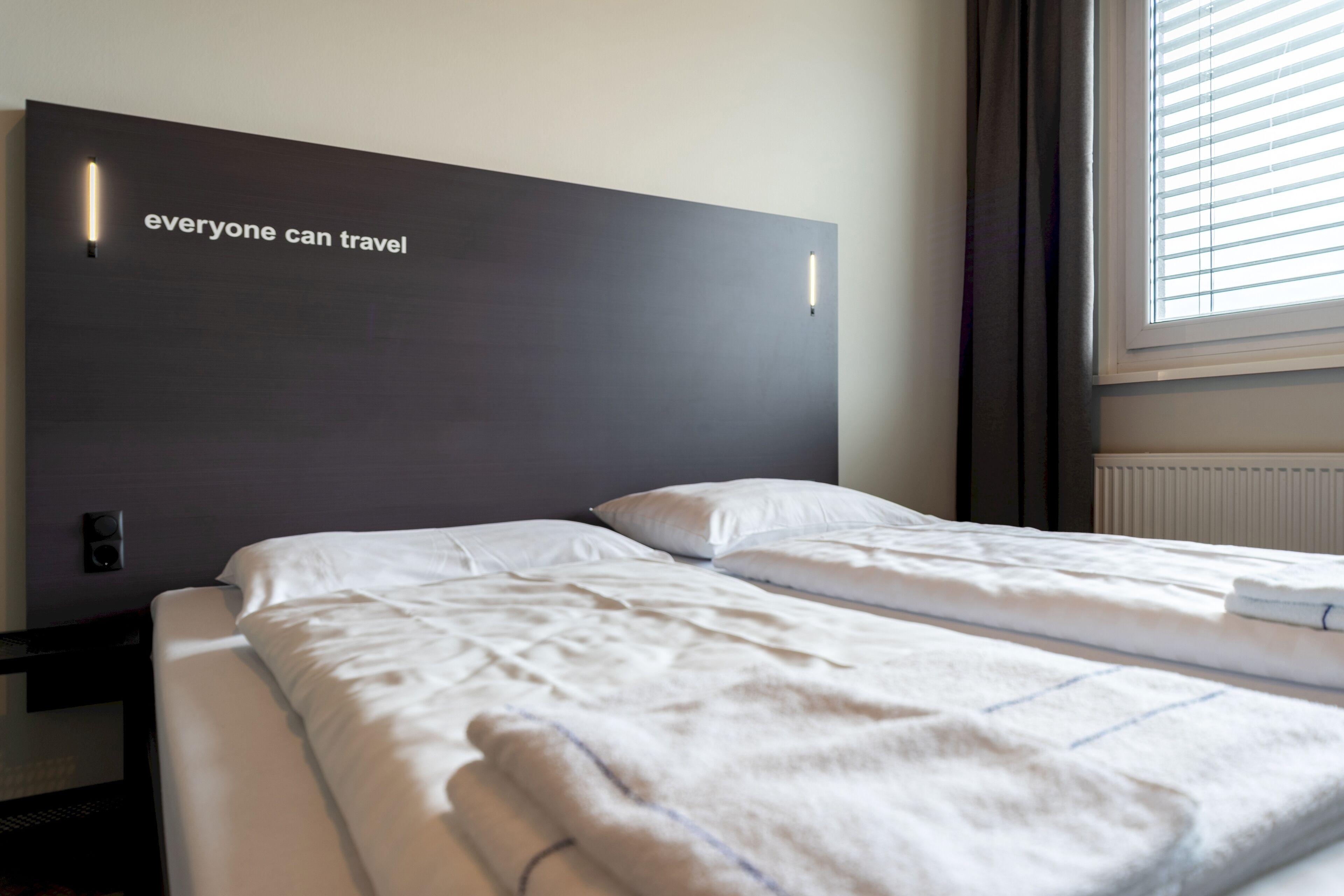 Ein Hotelzimmer mit einem großen Bett, einem Kopfteil mit einem Motivationsspruch, einem Fenster mit Jalousien und einem Nachttisch mit Lampe.