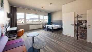 Ein modernes Hotelzimmer mit einem Bett, einem kleinen Tisch, einer Couch und Blick auf die Stadt durch große Fenster.