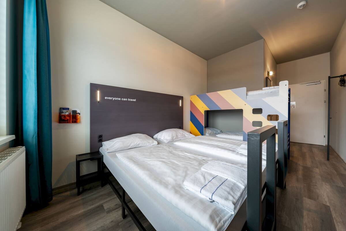 A hotel room with a bunk bed, a single bed, a wall-mounted TV, a radiator, and a window with curtains.