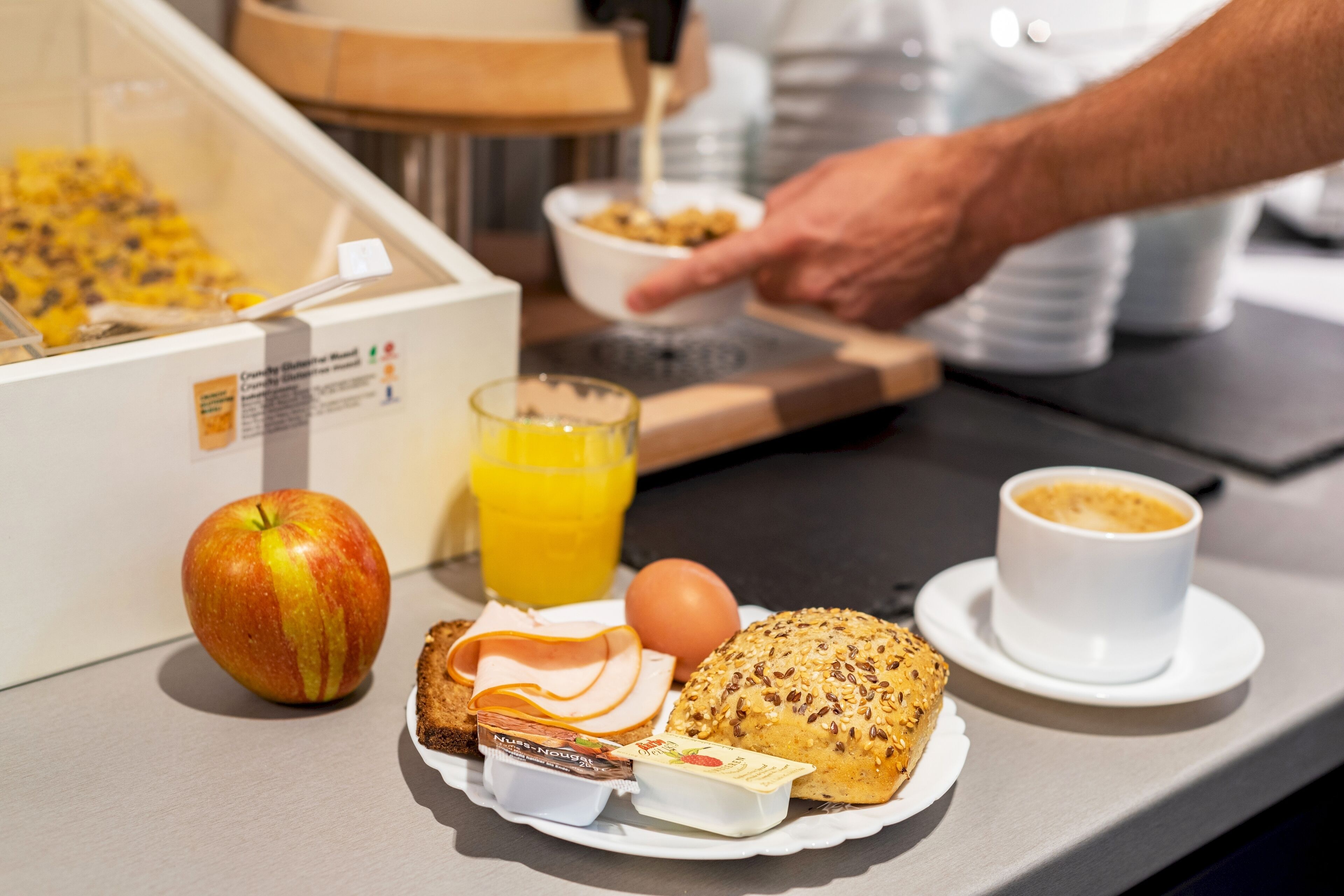 Une table mise pour le petit-déjeuner avec une assiette contenant une pomme, un verre de jus d’orange, une tasse de café, une tranche de pain et un pot de yaourt.
