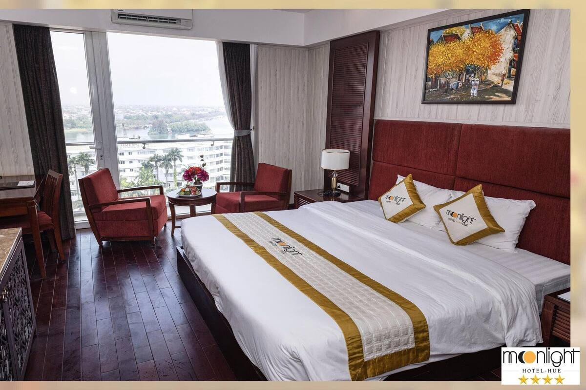 A hotel room with a large bed, a desk, a chair, and a view of the city through large windows.