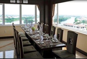 A dining room with a long table set for a meal, large windows with a view of a river and buildings, and modern chairs. - Moonlight Hotel Hue (Hue)