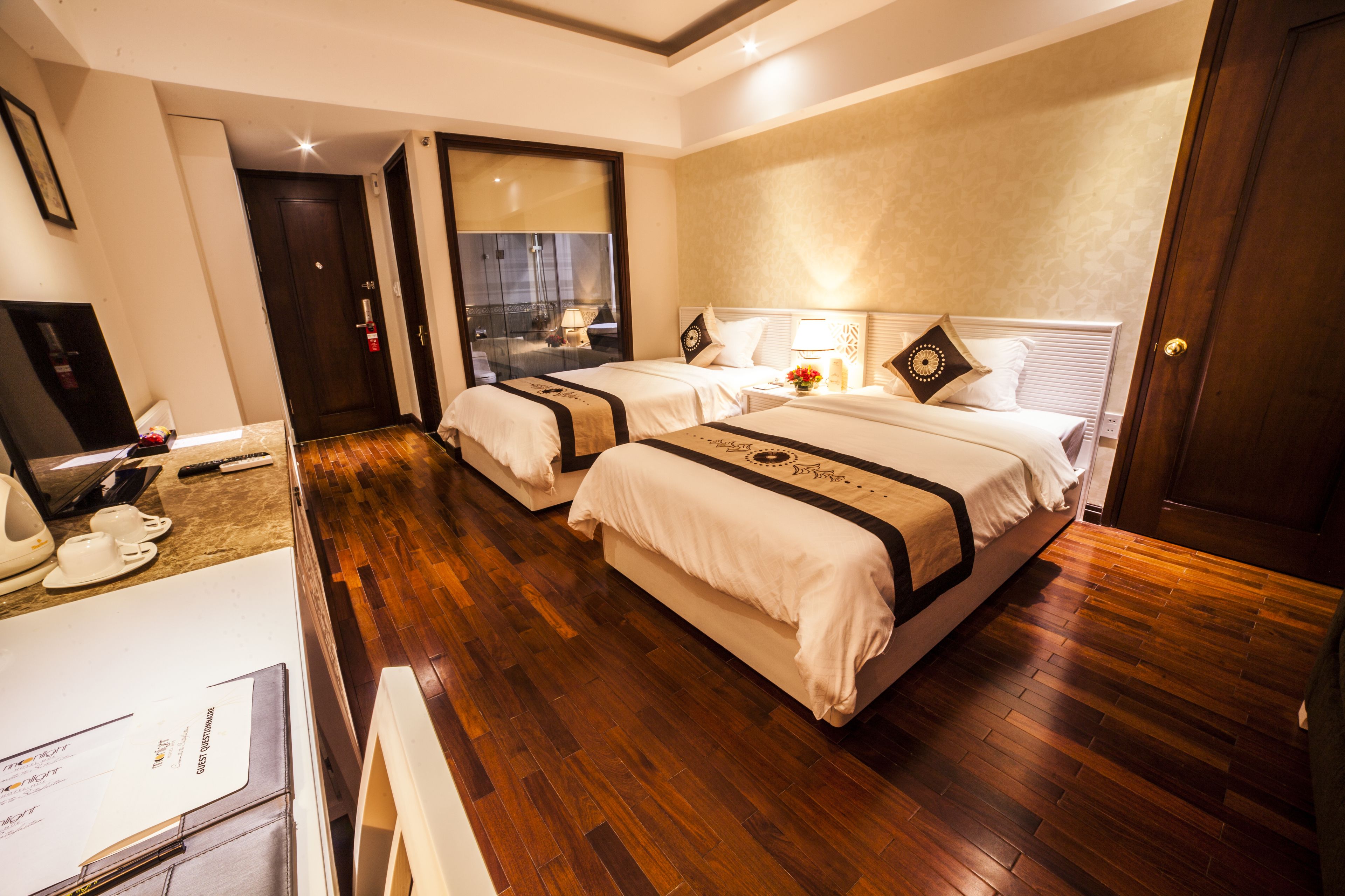Habitación de hotel con dos camas, suelo de madera, un escritorio con computadora y un baño con espejo y lavamanos.