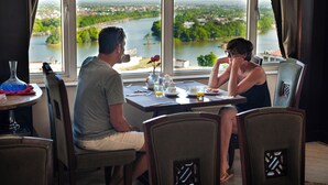 A dining area with a table set for two, a vase with a single flower, and a view of a river and greenery outside. - Moonlight Hotel Hue (Hue)