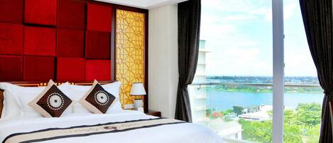 A hotel room with a large bed, a red headboard, a view of a body of water, and a balcony with curtains.