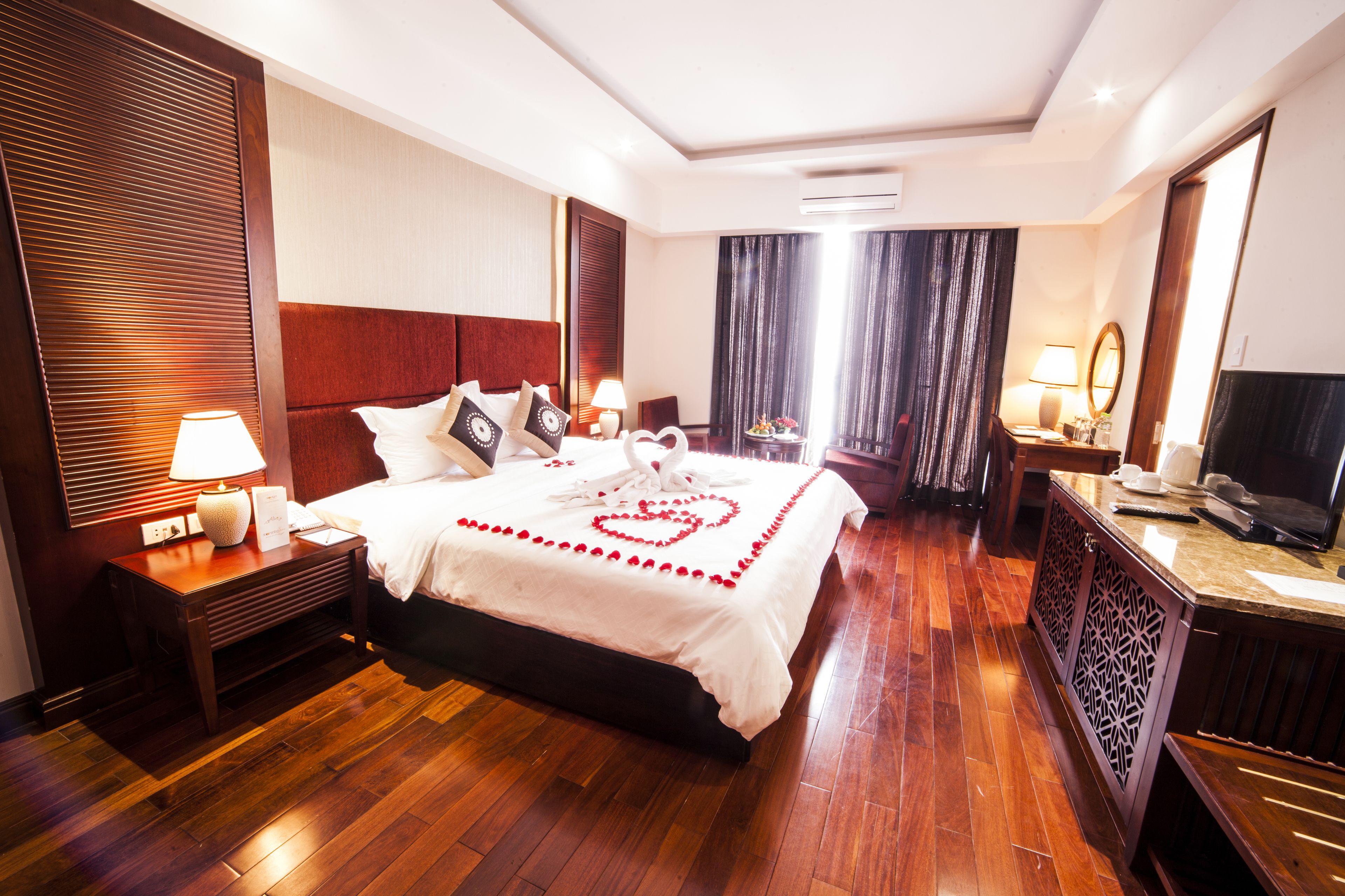 A hotel room with a large bed, wooden flooring, a nightstand with a lamp, a dresser with a mirror, and a television.