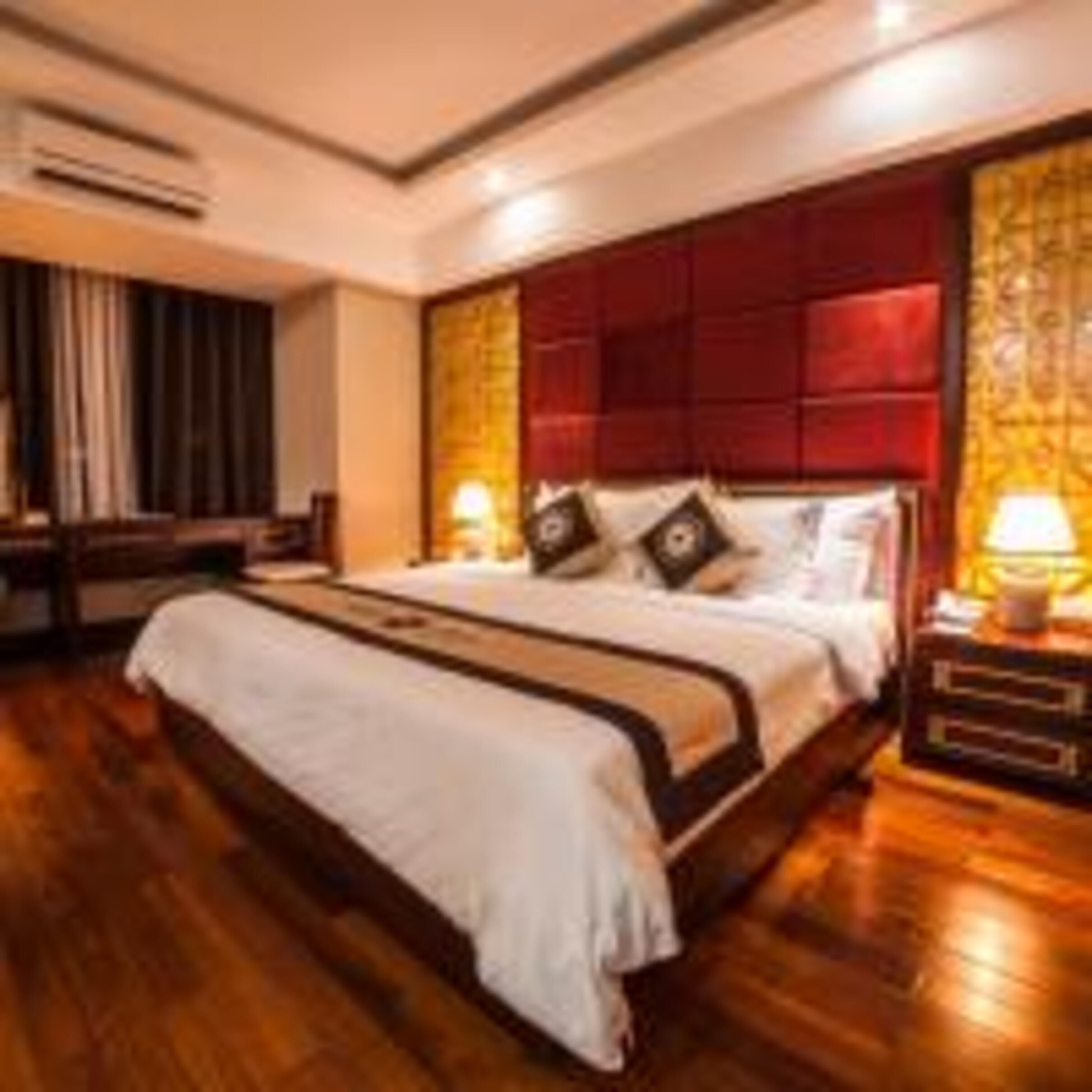 A hotel room with a large bed, wooden flooring, a red headboard, and a nightstand with a lamp.