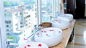 A hotel bathroom with three large, round sinks with rose petals, a wicker basket, and a view of the city through large windows. - Moonlight Hotel Hue (Hue)