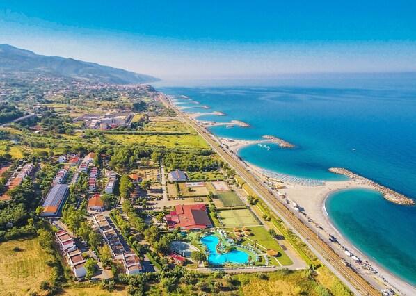 An aerial view of a coastal resort with multiple buildings, a swimming pool, and a beach area. - Villaggio Club Bahja (Paola)