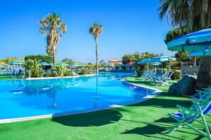 A swimming pool with blue chairs and umbrellas, surrounded by palm trees and greenery. - Villaggio Club Bahja (Paola)