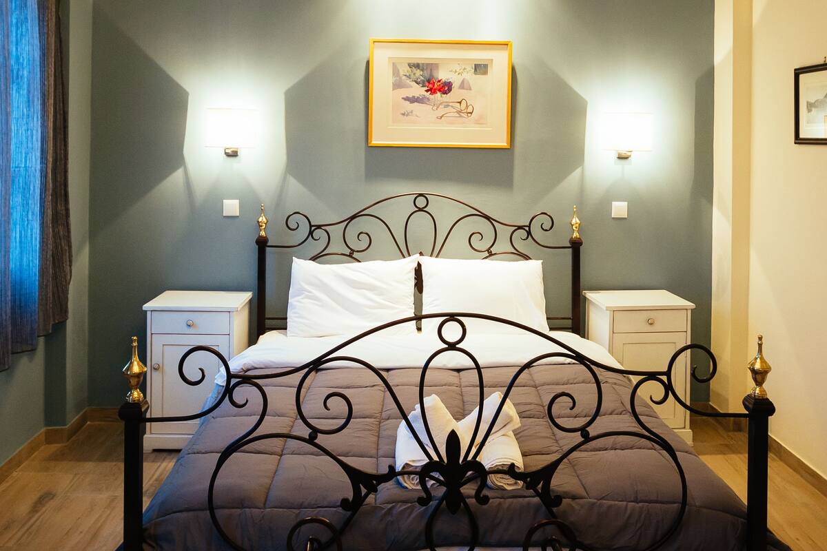 Un dormitorio con estructura de cama metálica, candelabro, lámparas de noche y un cuadro en la pared.