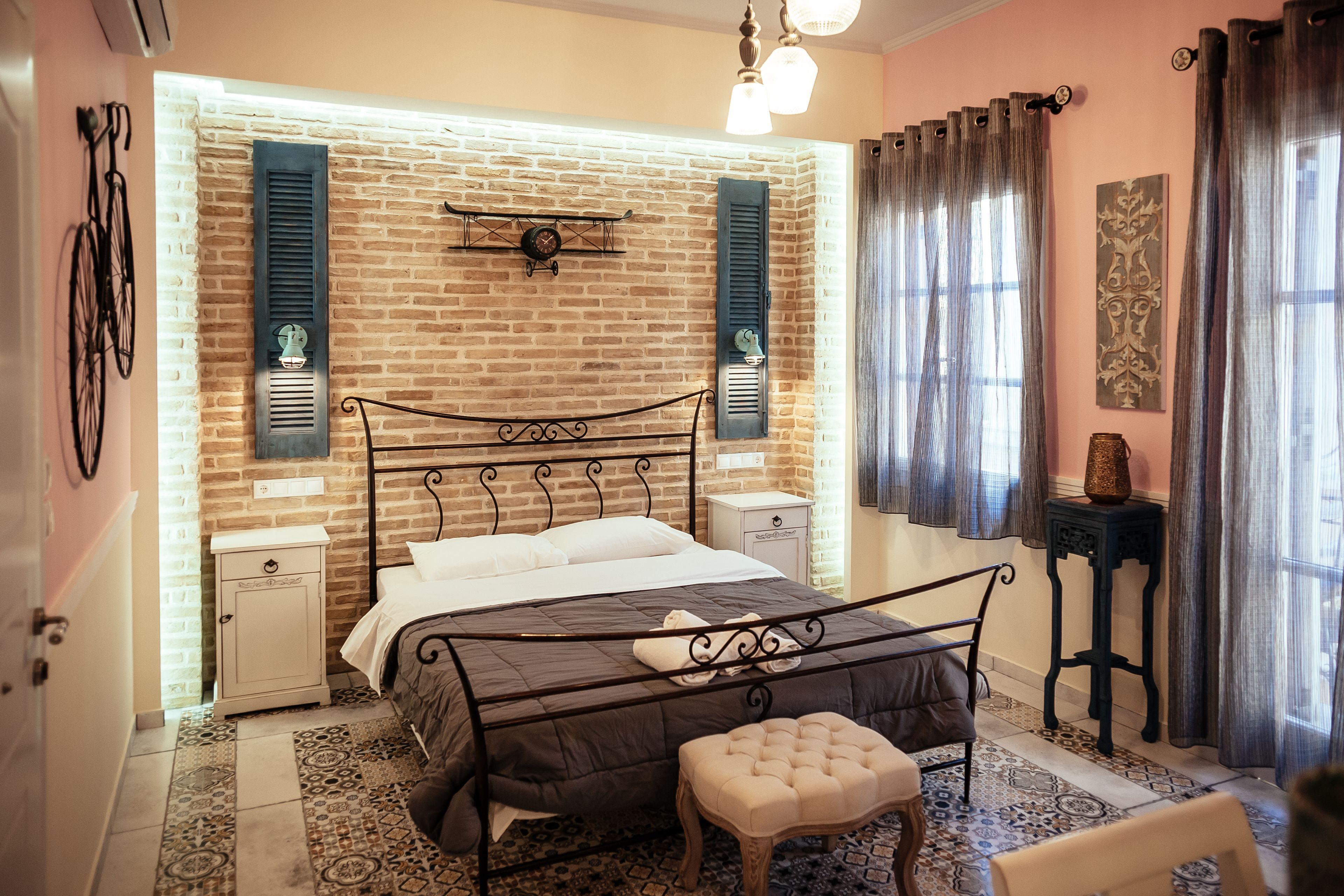 A bedroom with a brick wall, a bed with a headboard, bedside tables, a nightstand, a window with curtains, and a wall-mounted clock.
