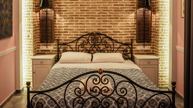 Un dormitorio con pared de ladrillo visto, cama con cabecera estampada, lámparas de noche, un banco y alfombra con diseño.