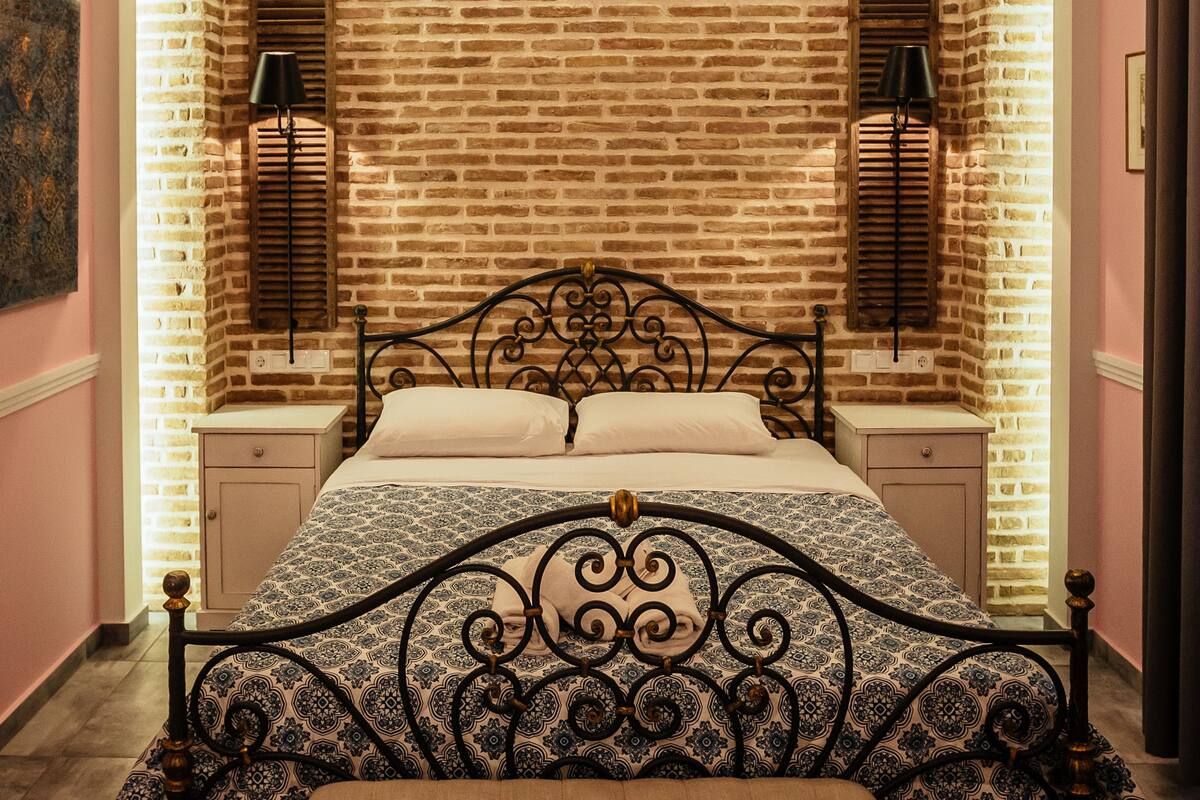 Un dormitorio con pared de ladrillo visto, cama con cabecera estampada, lámparas de noche, un banco y alfombra con diseño.