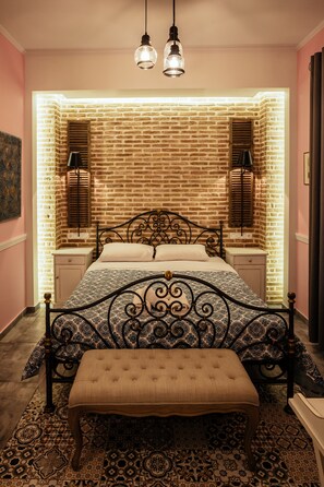 A bedroom with a brick-patterned wall, a bed with a patterned headboard, bedside lamps, a bench, and a patterned rug. - Pension Acronafplia (Nafplio)